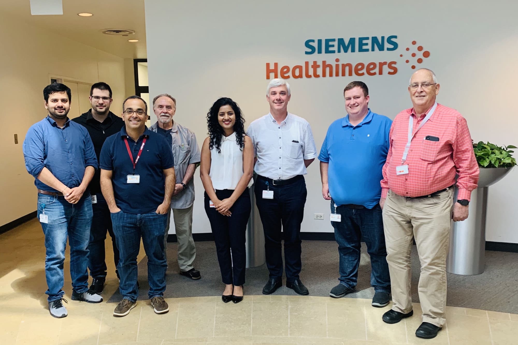 Harshitha with the SPECT R&D team at Siemens Healthineers, in front of the branded wall.